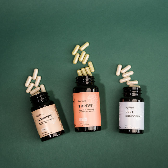 Three bottles of hey freya supplements lying on a forest green background with the capsules spilled out on the surface